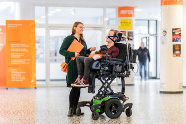 Foto eines Mitarbeiters im Rollstuhl, der auf Augenhöhe der Kollegin, die neben ihm steht, hochgefahren ist. Beide Kollegen unterhalten sich im Foyer. __ <br>Employee sits in a wheelchair, which has been raised at eye level with the colleague standing next to him. Both colleagues are talking in the foyer.