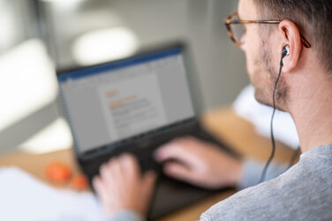 Foto eines Mannes am Laptop im Homeoffice mit Kopfhörern im Ohr. __ Man at laptop in home office with headphones in ear.