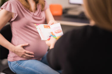 Foto einer schwangeren Frau, die einer Mitarbeiterin aus dem Familienservice gegenüber sitzt und einige Flyer zum Thema „Vereinbarkeit von Familie und Beruf“ entgegen nimmt.__Photo of a pregnant woman sitting across from a Family Service employee accepting some flyers on the topic of "Reconciling family and career."