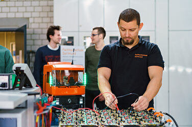 Labormitarbeiter misst etwas mit Messgeräten im Vordergrund. Dahinter stehen zwei Studierende, die sich unterhalten. Eine Lkw-Aufbaute leuchtet. __ Laboratory worker measures something with gauges in the foreground. Behind it are two students who are talking. A truck body lights up.