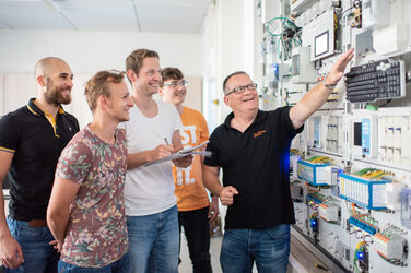 Foto eines Professors, der vor einer Versuchswand der Elektrotechnik steht und auf ein Gerät zeigt. Neben ihm stehen vier lächelnde Studenten, einer von ihnen macht sich Notizen. __ A professor stands in front of an electrical engineering test wall and points to a device. Next to him are four smiling students, one of whom is taking notes.