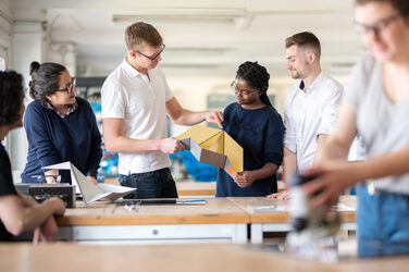 Photo of a lab member helping a student to fold a metal object. Next to them are other students watching the two. Another student in the foreground is working. __ <br>A laboratory worker helps a student to fold a metal object. There are other students standing around him and around the workbench, watching the two of them. Another student is out of focus in the foreground and is doing something.