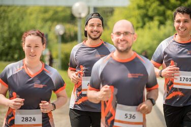 Foto von vier Mitarbeiter*innen in FH-Laufkleidung, die bei einem organisierten Lauf mitlaufen. __ Employees with FH equipment take part in an organized run.