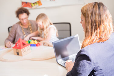 Foto aus einem Büro. Rechts im Bild arbeitet eine Frau am Laptop, während ihre kleine Tochter mit der Oma an einem Tisch mit einer Holzlokomotive spielt.__Photo from an office. On the right side of the picture a woman is working on a laptop while her little daughter is playing with a wooden locomotive at a table with her grandma.