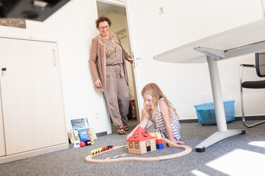 Photo of a little girl playing with a wooden train set on the floor of an office. Her grandma walks in the door and smiles down at her.__Photo of a little girl playing with a wooden train set on the floor of an office. Her grandma walks in the door and smiles down at her.