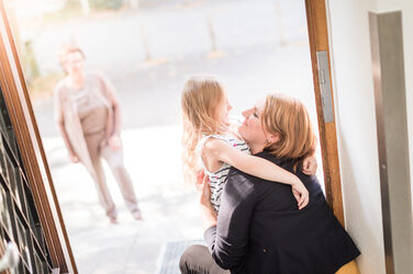 Photo of a young woman squatting and hugging her little daughter. In the background you can see the grandma.__Photo of a young woman squatting and hugging her little daughter. In the background you can see the grandma.