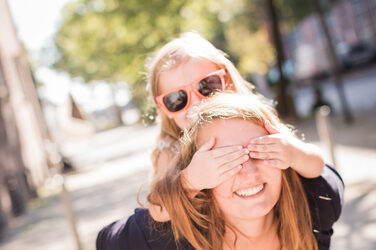 Foto einer junge Frau, die ein kleines Mädchen mit Sonnenbrille huckepack trägt. Das Mädchen hält der Frau die Augen zu.__Photo of a young woman piggybacking a little girl with sunglasses. The girl holds the woman's eyes.