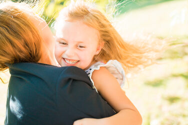 Foto einer Frau, von der man das Gesicht nicht sehen kann, mir ihrer kleinen Tochter auf dem Arm, welche freudig lacht.__Photo of a woman, whose face cannot be seen, with her little daughter in her arms, laughing joyfully.