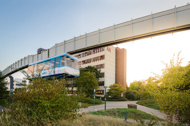 Photo of the H-Bahn tracks with passing H-Bahn, in the background the building Emil-Figge-Straße 44 of the Fachhochschule Dortmund.