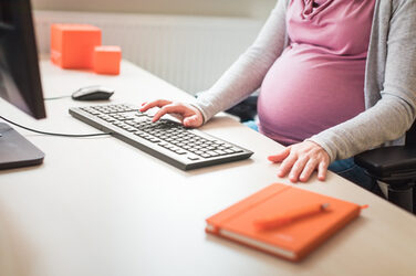 Photo of pregnant employee sitting at desk and working on PC. Her head is not included in the picture __Photo of pregnant employee sitting at desk and working on PC. Her head is not included in the picture.
