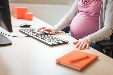 Foto einer schwangeren Mitarbeiterin, die am Schreibtisch sitzt und am PC arbeitet. Ihr Kopf ist nicht mit auf dem Bild.__Photo of pregnant employee sitting at desk and working on PC. Her head is not included in the picture.