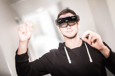 Foto von einem Studenten, der eine VR-Brille trägt und die Hände zur Steuerung in die Höhe hält.__ Student wears VR glasses and holds hands up to control.