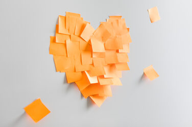 Foto von einem aus orangenen Haftnotizzetteln zusammengeklebten Herz an einer Wand. __ A heart stuck together from orange sticky notes on the wall.