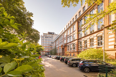 Foto des Altbaus und des Hochhauses an der Sonnenstraße mit Parkplatz davor. Links im Bild im Anschnitt ein blühender Busch.