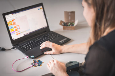 Foto einer Studentin am Laptop. Per USB-Kabel ist ein Calliope - ein Mikrocontroller - an den Laptop angeschlossen.