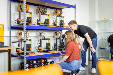 Photo of two students standing/squatting at the 3D printer shelf.