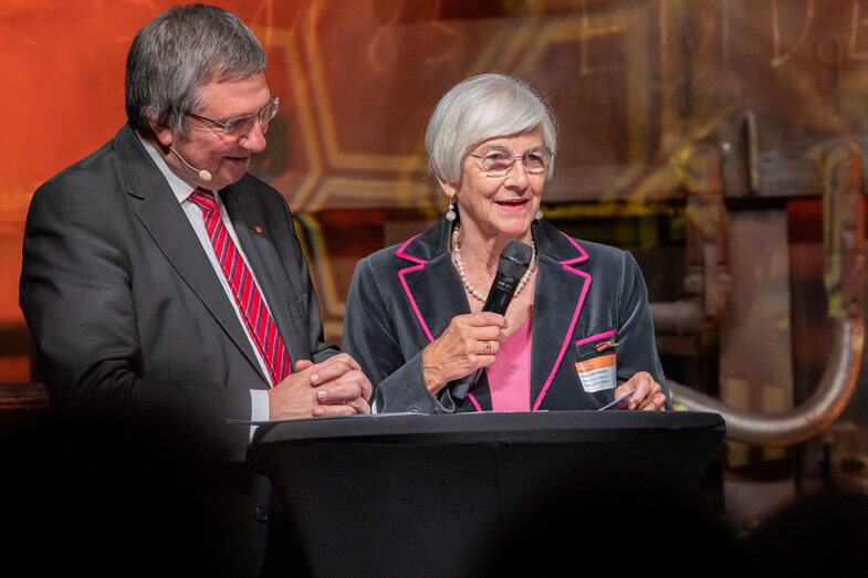 Photo of a male and a female person. The female person speaks into a microphone.