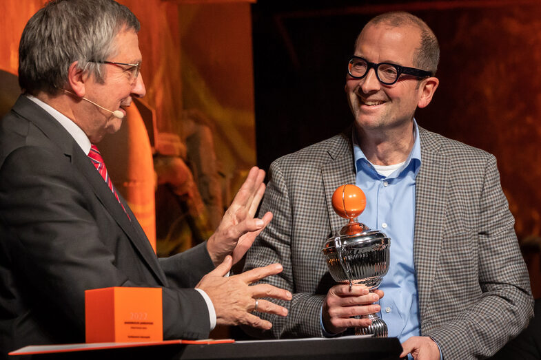 Photo of two men talking to each other. One person is holding a cup in his hand.