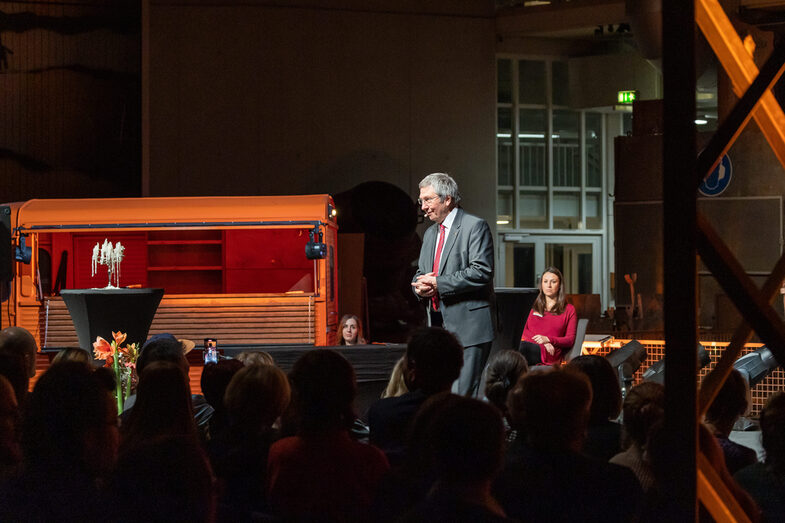 Photo of a male person. This person stands on a stage and speaks to people sitting in front of him in the audience.