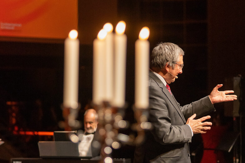 Photo of a male person speaking into a microphone. In the foreground is a candlestick with five burning candles on a high table. In the background, a male person is sitting in front of an electric piano.