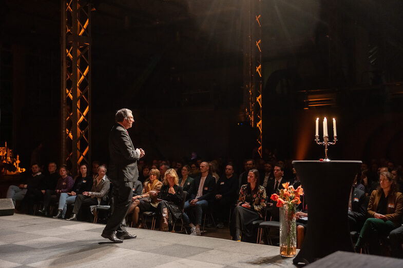 Photo of a male person. This person stands on a stage and speaks to people sitting in front of him in the audience.