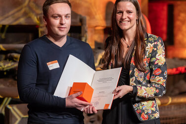 Foto von einer männlichen und einer weiblichen Person die stehend in die Kamera lächeln. Die männliche Person hält einen Würfel und eine Urkunde in der Hand.