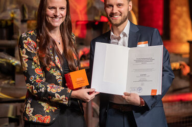Foto von einer männlichen und einer weiblichen Person die stehend in die Kamera lächeln. Die männliche Person hält einen Würfel und eine Urkunde in der Hand.
