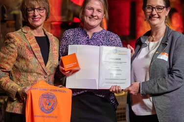 Foto von drei weiblichen Personen die stehend in die Kamera lächeln. Eine Person hält eine orangene Weste in der Hand. Eine zweite Person hält einen Würfel und eine Urkunde in der Hand.