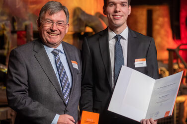 Foto von zwei männlichen Personen die stehend in die Kamera lächeln. Eine Person hält einen Würfel in der Hand. Die andere Person hält eine Urkunde in der Hand.