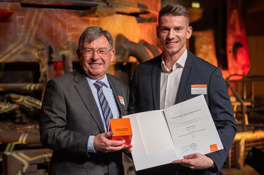 Foto von zwei männlichen Personen die stehend in die Kamera lächeln. Eine Person hält einen Würfel in der Hand. Die andere Person hält eine Urkunde in der Hand.