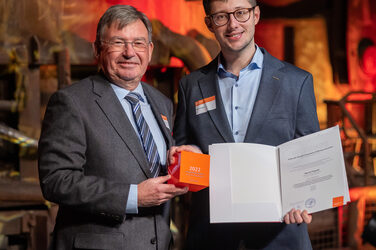 Foto von zwei männlichen Personen die stehend in die Kamera lächeln. Eine Person hält einen Würfel in der Hand. Die andere Person hält eine Urkunde in der Hand.