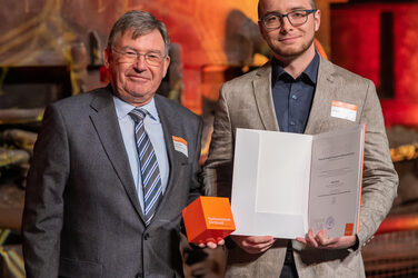 Foto von zwei männlichen Personen die stehend in die Kamera lächeln. Eine Person hält einen Würfel in der Hand. Die andere Person hält eine Urkunde in der Hand.