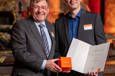 Foto von zwei männlichen Personen die stehend in die Kamera lächeln. Eine Person hält einen Würfel in der Hand. Die andere Person hält eine Urkunde in der Hand.