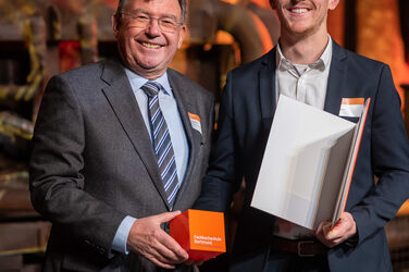 Foto von zwei männlichen Personen die stehend in die Kamera lächeln. Eine Person hält einen Würfel in der Hand. Die andere Person hält eine Urkunde in der Hand.