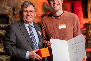 Foto von zwei männlichen Personen die stehend in die Kamera lächeln. Eine Person hält einen Würfel in der Hand. Die andere Person hält eine Urkunde in der Hand.