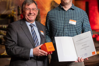 Foto von zwei männlichen Personen die stehend in die Kamera lächeln. Eine Person hält einen Würfel in der Hand. Die andere Person hält eine Urkunde in der Hand.