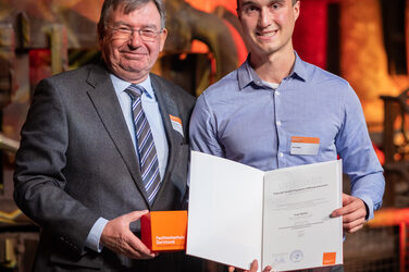 Foto von zwei männlichen Personen die stehend in die Kamera lächeln. Eine Person hält einen Würfel in der Hand. Die andere Person hält eine Urkunde in der Hand.