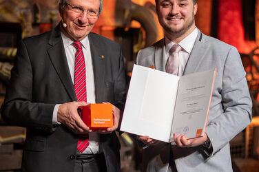 Foto von zwei männlichen Personen die stehend in die Kamera lächeln. Eine Person hält einen Würfel in der Hand. Die andere Person hält eine Urkunde in der Hand.