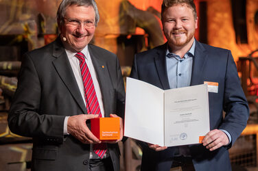Foto von zwei männlichen Personen die stehend in die Kamera lächeln. Eine Person hält einen Würfel in der Hand. Die andere Person hält eine Urkunde in der Hand.