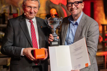 Foto von zwei männlichen Personen die stehend in die Kamera lächeln. Eine Person hält einen Würfel in der Hand. Die andere Person hält eine Urkunde und einen Pokal in der Hand.