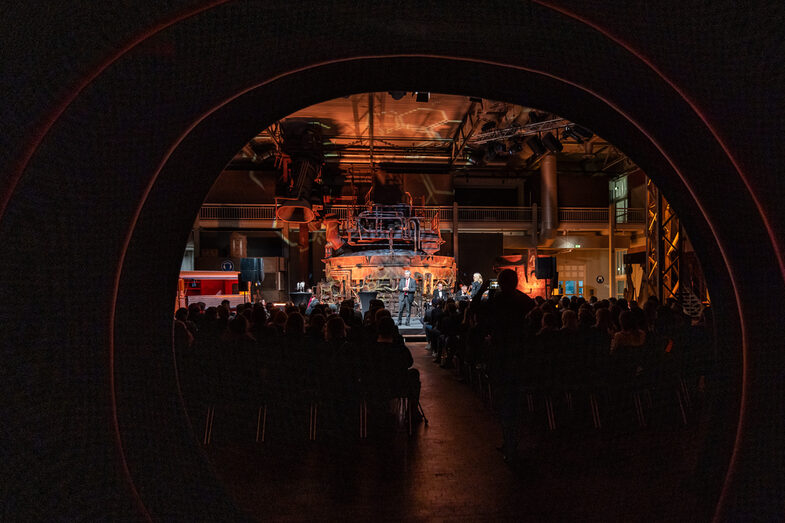 Photo of an illuminated hall. In it, a male person is standing on a stage and speaking to the audience sitting in front of the stage.