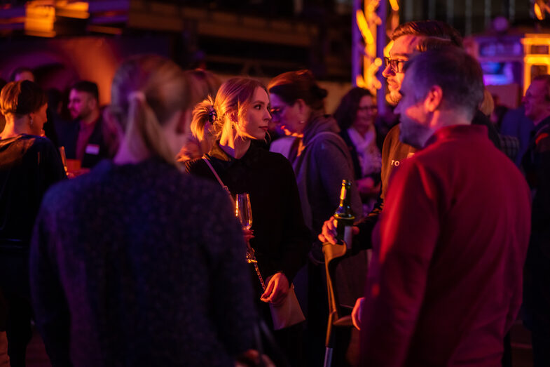 Several male and female people are standing in an illuminated hall and talking.