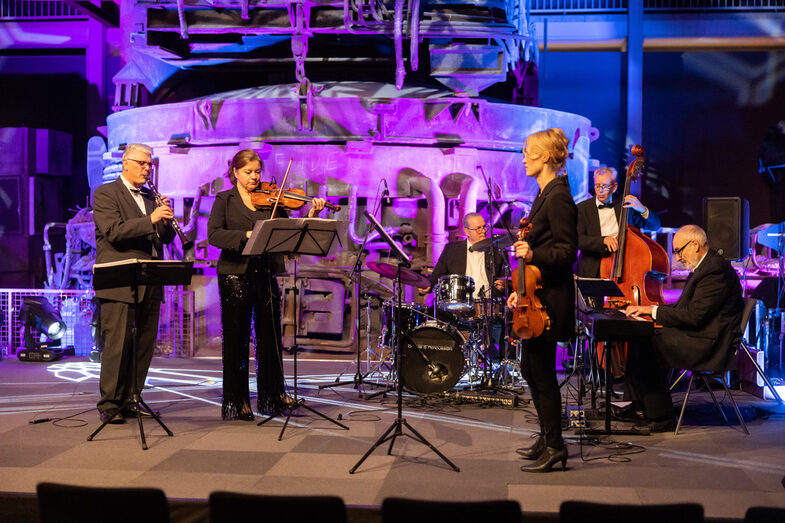 Photo of a band playing on a stage. The band consists of two females and four males. Each person plays a musical instrument.