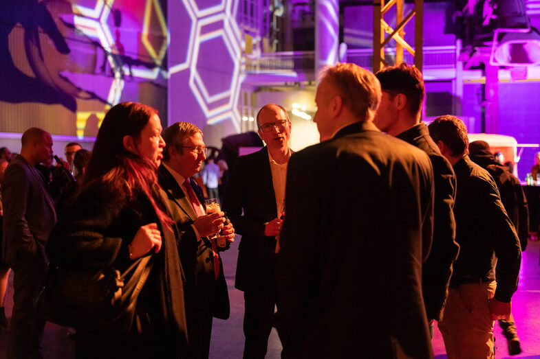 Several male and female people are standing in an illuminated hall and talking.