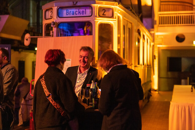 Photo of a male and two female persons. They are standing at a bar table in front of an old streetcar and talking.