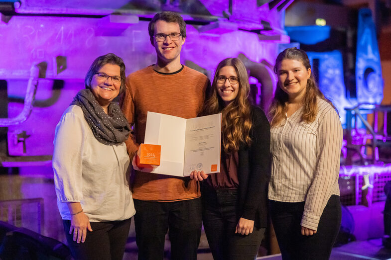 Photo of one male and three female people smiling at the camera. The male person is holding a certificate and a dice.