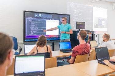Photo of a lecture situation in a seminar room. A man is standing in front of a digital board and explaining something in code.