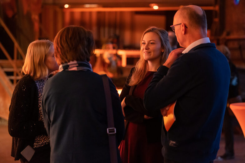 Photo of three female and one male person standing in a hall and talking.