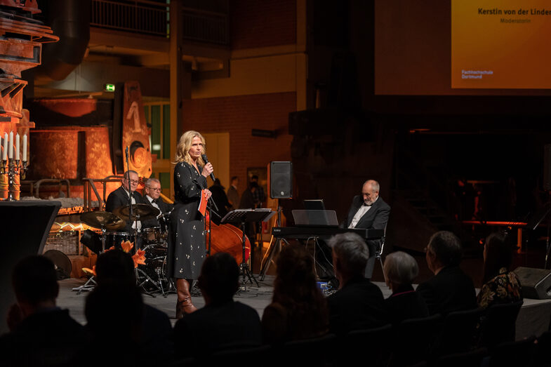 Photo of a female person. She is standing on a stage and speaking into a microphone to the audience sitting in front of the stage. In the background of the stage, three male members of a band are sitting in front of their musical instruments.