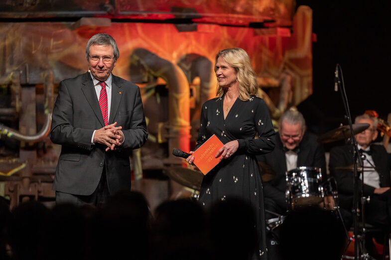 Photo of a male and a female person on a stage. The male person is speaking into a microphone in front of an audience.